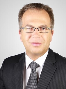Borisenko mit Brille, Anzug und Krawatte, frontal aufgenommen vor grauem Hintergrund.